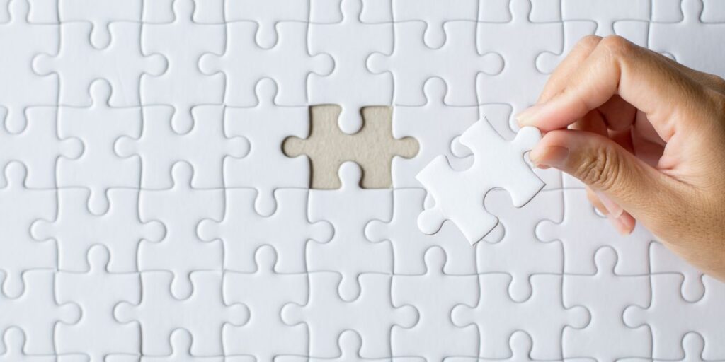 Hand placing a missing piece into a white jigsaw puzzle.