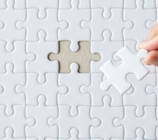Hand placing a missing piece into a white jigsaw puzzle.
