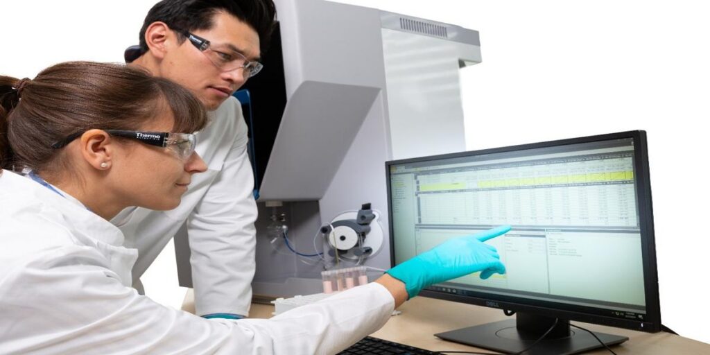 Two scientists in lab coats reviewing analytical data on a computer beside lab instrumentation.