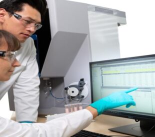Two scientists in lab coats reviewing analytical data on a computer beside lab instrumentation.