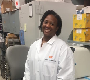 Dr. Athena Starlard-Davenport, a Black researcher in a lab coat, smiles in her lab, where she researches breast cancer