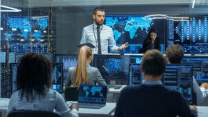 person standing at front of network operations center talking to colleagues surrounded by computers and monitors displaying global systems security status