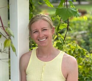 Portrait of a smiling plant pathology researcher standing outdoors beside grapevine leaves.
