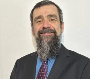 Headshot of a man with dark hair and a beard wearing a suit and tie against a neutral background.