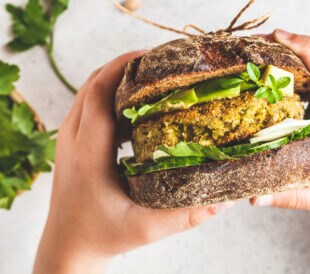 Vegan sandwich with chickpea patty, avocado, cucumber and greens in rye bread in children's hands, top view.