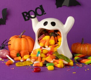 Festive Halloween side view tabletop scene featuring a ghost bucket filled with colorful candy, pumpkins, and bats on purple background. Perfect for creating a spooky and celebratory Halloween design