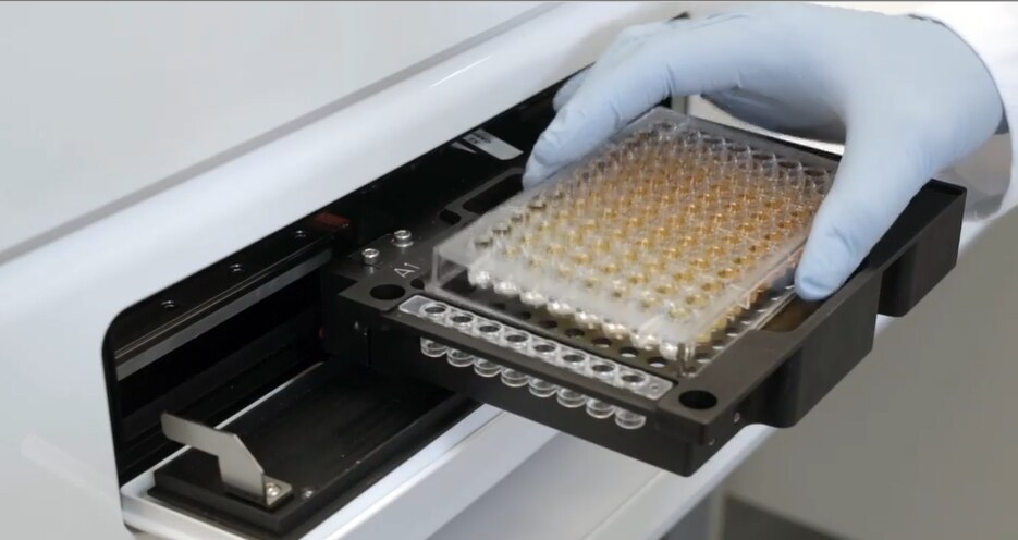 A scientist in PPE inserts a 96 well plate into a multimode microplate reader
