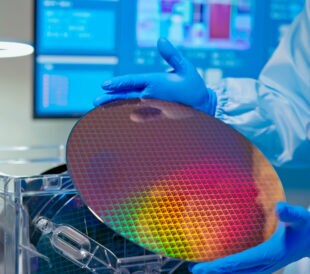 close up of asian male technician in sterile coverall holds wafer that reflects many different colors with gloves and check it at semiconductor manufacturing plant