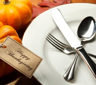 A tag with "Happy Thanksgiving" printed on it is tied to a small pumpkin that isits next to a dinner plate set for a Thanksgiving dinner.