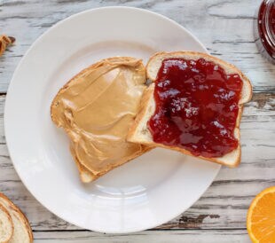 Top view of open face homemade peanut butter and strawberry Jelly sandwich on oat bread, over a white rustic wooden table / background. Served with fresh oranges / fruit.