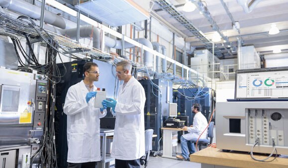 两名戴着护目镜、身穿实验室白色工作服的男科学家在锂离子电池测试设施中谈到: 两名戴着护目镜、身穿实验室白色工作服的男科学家在锂离子电池测试设施中谈到: